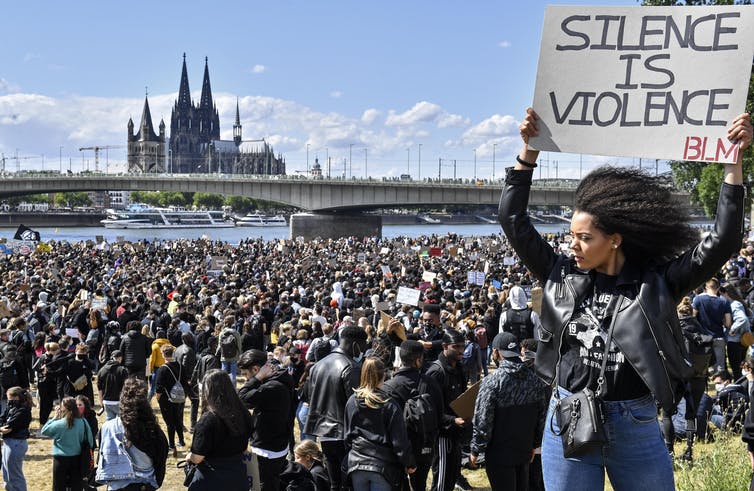 A woman holding a sign reading 'Silence is violence' and 'BLM' stands in front of a crowd