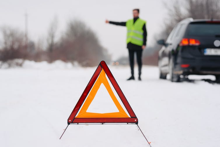 A man in a hi-vis vest stands by his car on the road side in the snow, with a hazard triangle.