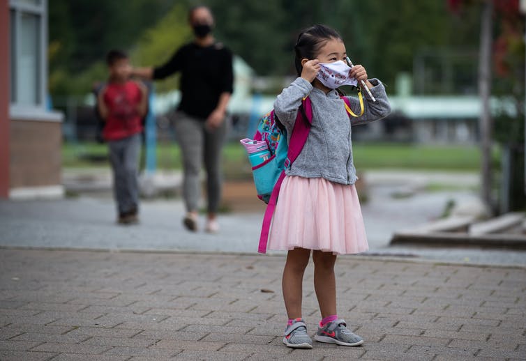 A little girl in a pink skirt and grey jacket with a turquoise and pink backpack puts on her face mask.