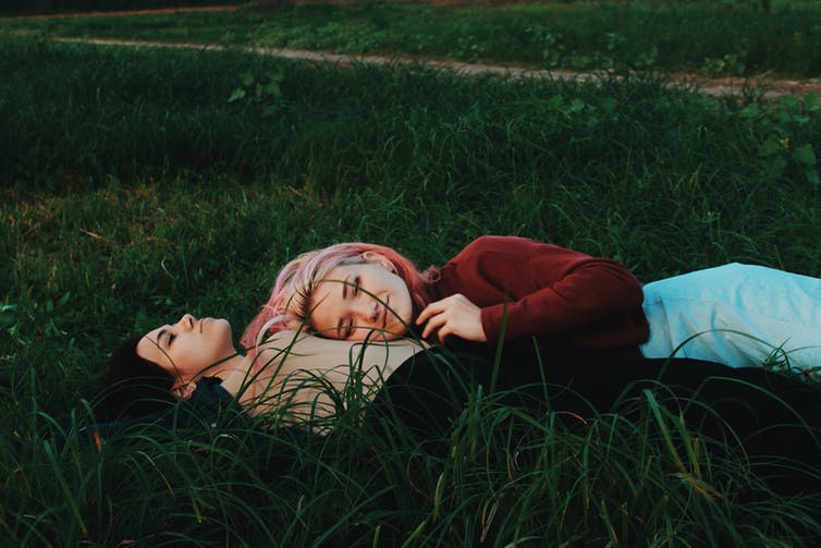 Two teenaged girls lay among grass, cuddling.