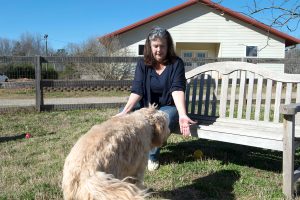 Jennifer Arnold sits on a bench with her arm extended as Great, a fluffy goldendoodle, approaches to tap her hand..