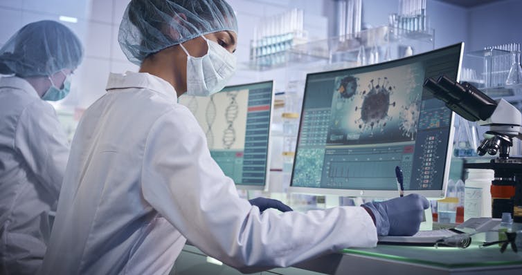 Two young scientists wearing protective masks and caps working on their computers that have an image of the coronavirus.