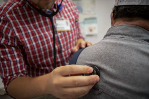 A man wearing a red button down shirt presses an instrument to a man's back.