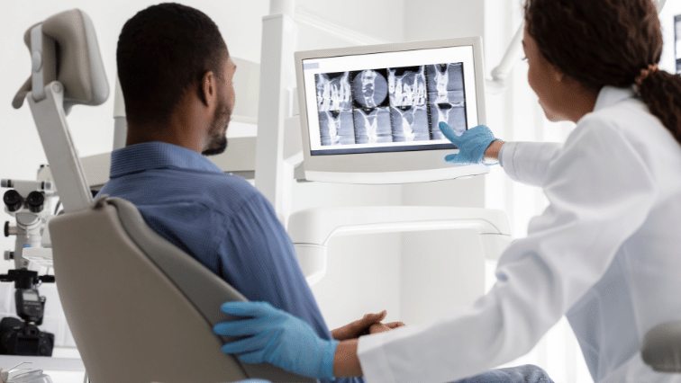 An image of a black patient, seated in a large operating chair, and a Black doctor standing next to him and pointing to an series of medical scan images that are displaying on a computer screen above them both.