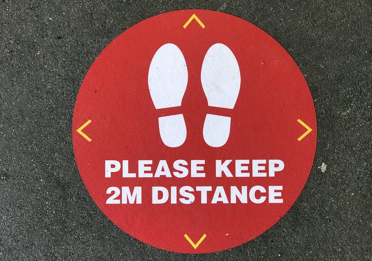 Close-up image of a red social distancing circle on asphalt, with two shoe-prints and the message 'Please keep 2M distance'