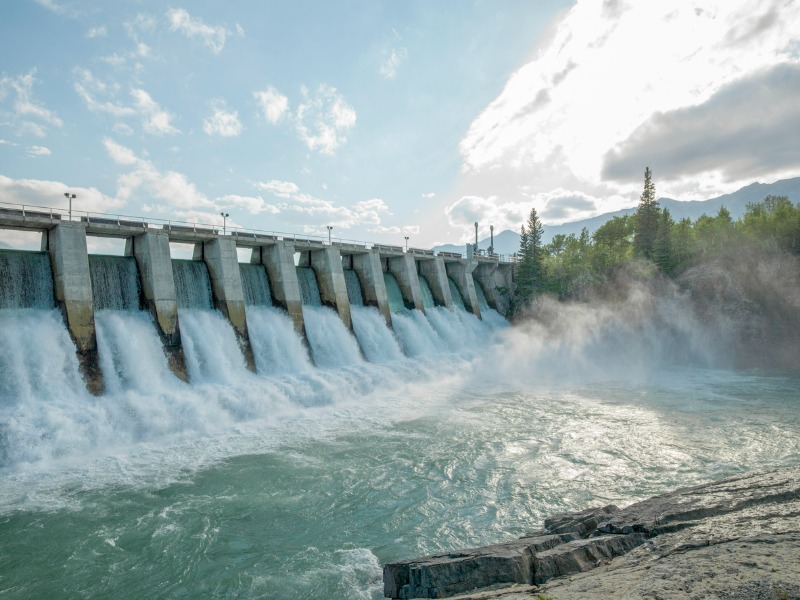 Water rushes through hydroelectric dam. A forest and mountains can be seen in the distance.