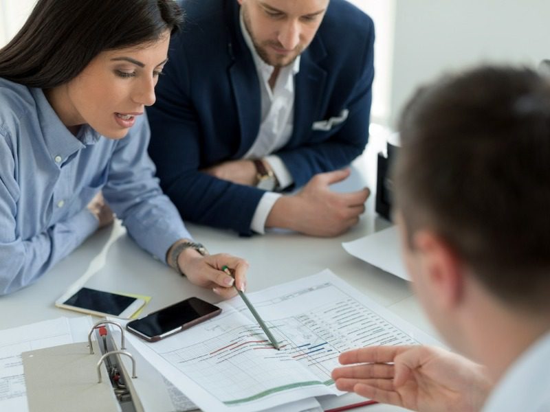 A woman and a man sit across from another man at a desk. The woman uses a pen to point at a stack of papers. They are all dressed in business attire.