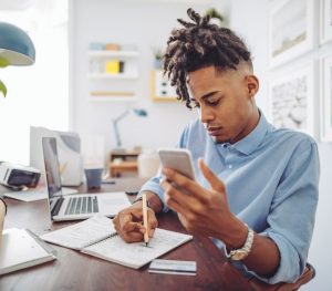 A young man sits at a desk. He is writing on a notepad with one hand and is scrolling on his phone in the other. He sits in front of a desk with a laptop.