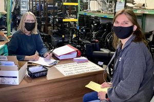 two women in face masks sit at a desk, wheelchairs are stacked behind them