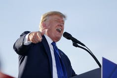 A blond, red-faced man yells into a microphone while pointing.