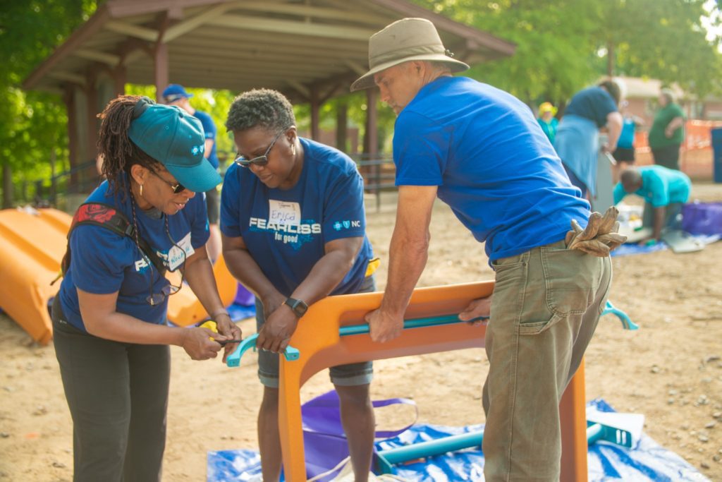 Three Blue Cross NC volunteers in matching blue shirts work together to assemble a piece of children's play equipment