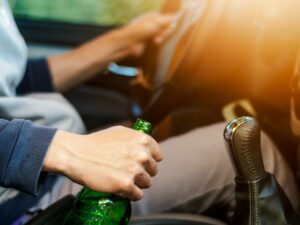 a man holds a green beer bottle in one hand and the steering wheel of his car in the other