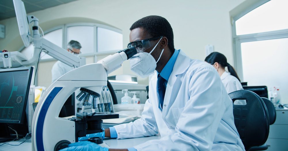 Black male pathologist in gloves and a mask looks through a microscope
