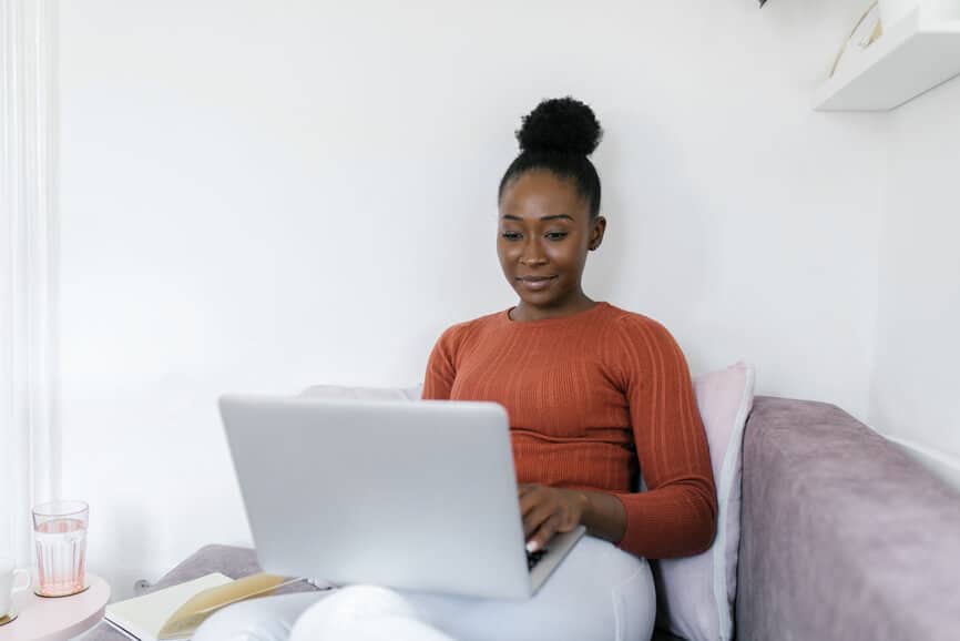 Beautiful African American woman working from home. She is sitting on the sofa in a cozy living room with laptop in her lap.