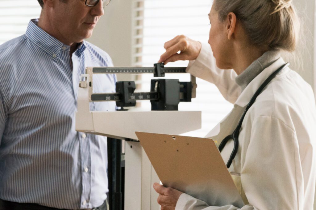 A photo shows a male patient being weighed on a scale by a doctor.