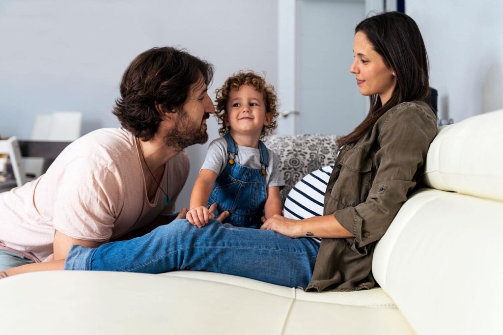 Pregnant mom and dad sitting on a bed with their toddler.