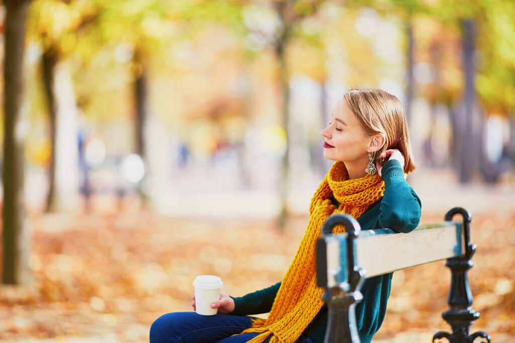 A woman sits on a park bench, enjoying her coffee and considering things with her eyes closed