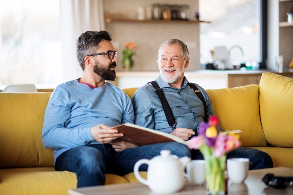 An adult son and senior father sitting on sofa indoors at home, talking.