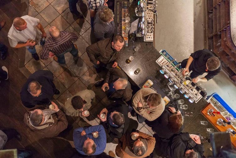 From above we see the heads of a group of customers as they mingle around a bar holding pints of beer in their hands.