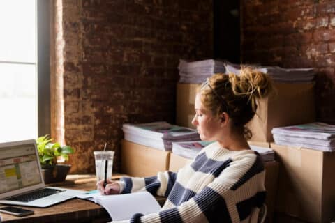 Woman working at desk, writing in notebook in front of computer at publishing company next to stacks of newspapers.