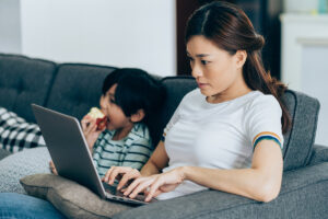 A woman researches her insurance plan on the couch, as her son eats an apple alongside.