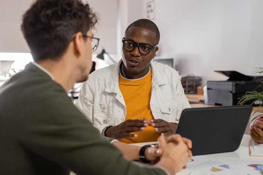 African American guy in casual clothes gesticulating and explaining project data to male coworker while sitting at table in modern workspace