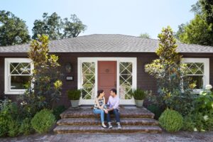 Happy couple sitting on the front steps of the new home they purchased together as their first home.