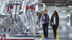 Female engineers discussing by car chassis. Full length of professionals are walking on aisle in factory. Colleagues are in automobile industry.