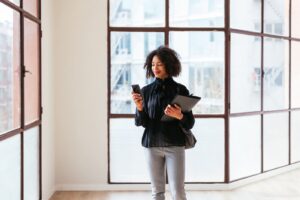 Glad black woman with curly hair smiling and browsing mobile phone while selling spacious light apartment