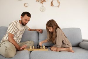 Bearded father watching smart kid making move while sitting on couch and teaching daughter to play chess on weekend day in daytime at home.