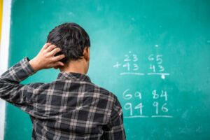 Confused kid scratching head to solve mathematics problem on board at classroom - concept of intelligence, trouble learning and education.