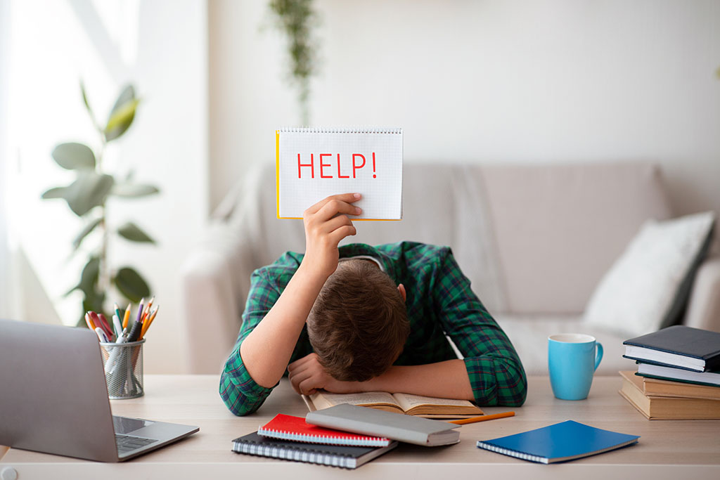 Man or boy at his desk, leaning his head on a pile of books in front of his laptop with a note on a notepad saying "help" 