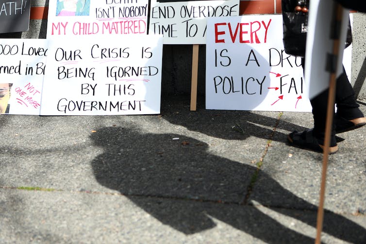 A person's shadow in the foreground in front of protest signs leaning on the ground, criticizing government inaction on the overdose crisis