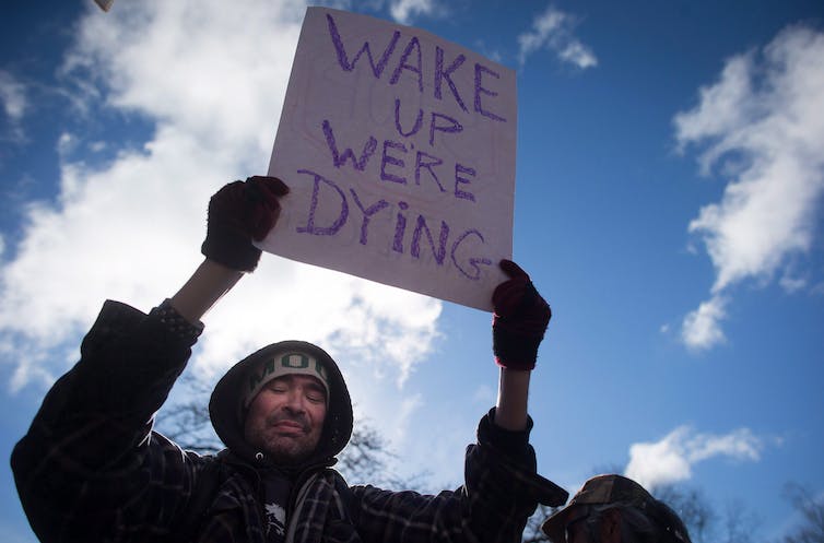 A man in a hoodie holds a sign over his head reading 'Wake up we're dying'