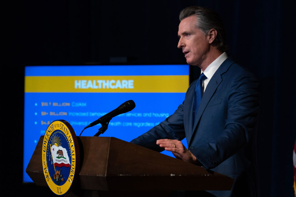 A photo of Governor Gavin Newsom speaking at a podium indoors with a presentation about health care seen on a screen behind him.