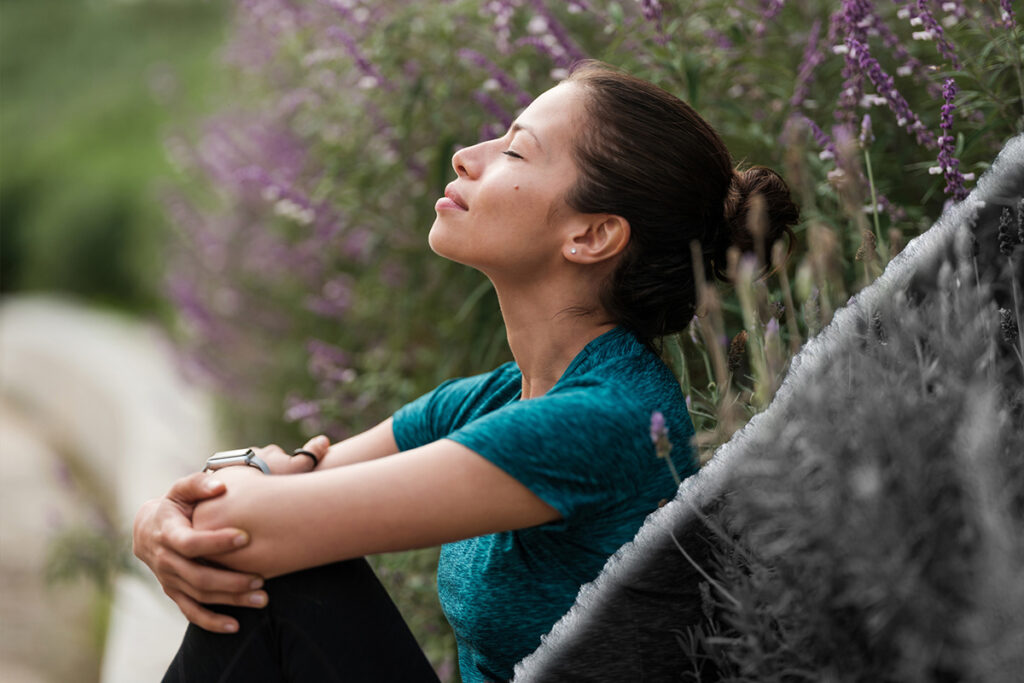 A woman sitting with her eyes closed and relaxing next to some flowers.