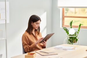 Focused trendy lady in casual clothes sitting at table and using tablet while working on startup project in light home office