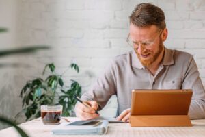 Portrait of man with glasses sitting in office and working, using digital tablet and writing something