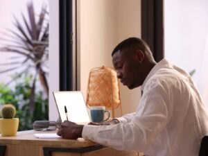 Young black man in white shirt writing memos on paper while sitting at table with netbook and doing freelance work