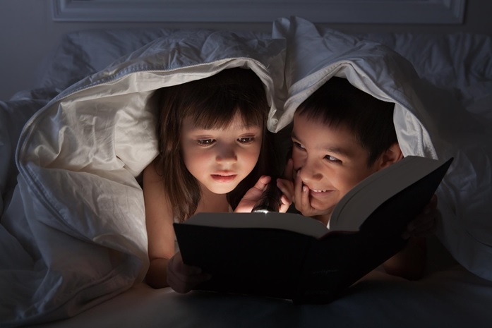 A boy and a girl reading a book with a torch under the blanket