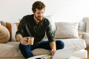 Caucasian middle age man sitting, holding cup of coffee and looking at his laptop placed on coffee table in the living room.