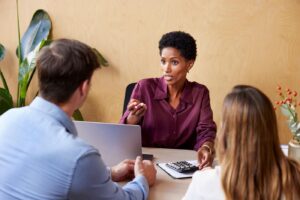 African American woman with calculator sitting at table and giving financial adviser to clients during meeting in office.