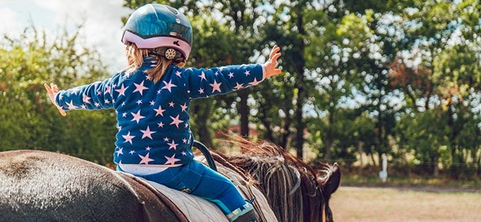 Little girl facing away from the camera with her arms outstretched while she sits on horseback. She's wearing a helmet and a blue fleece with pink stars. The setting is in a field surrounded by woods.