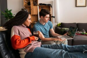 Contemporary interracial couple relaxing at home in their loft, drinking coffee and browsing social media on laptop