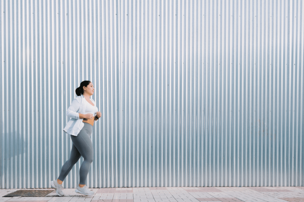 A woman is running in the street. She is in front of a great metallic wall that shines with the sunlight of the sunset. There is copy space.