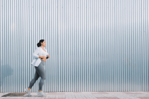 A woman is running in the street. She is in front of a great metallic wall that shines with the sunlight of the sunset. There is copy space.