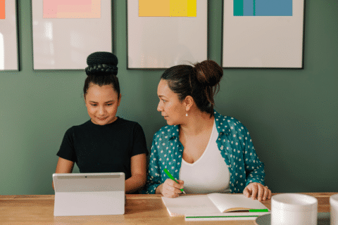 Woman looking at a girl while she works in a computer device sitting on a table at home