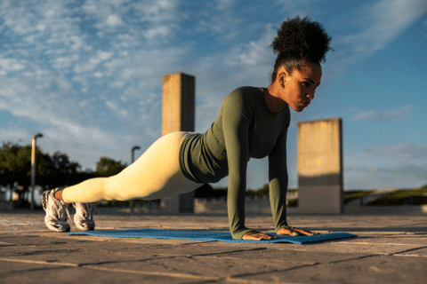 Hispanic sporty woman with afro doing core workout and abdominal planks wearing sportswear in urban scenery at sundown