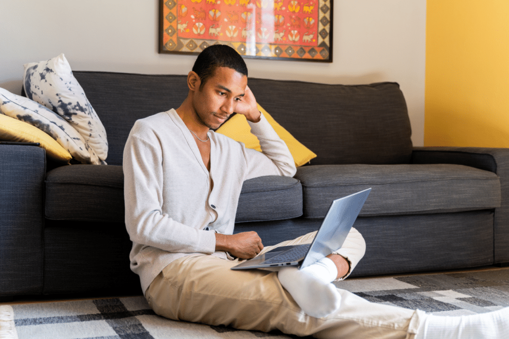 Happy young man working from home with laptop doing videocall sitting on couch at living room at home during daytime