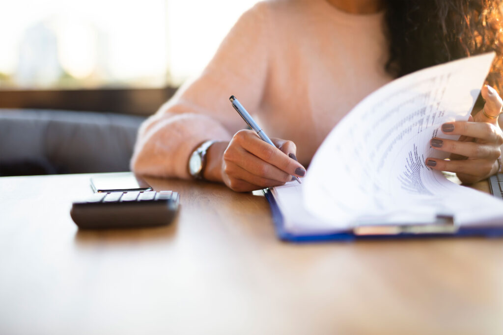 A person sits at a table with a phone, calculator and writes on paperwork on a clipboard.